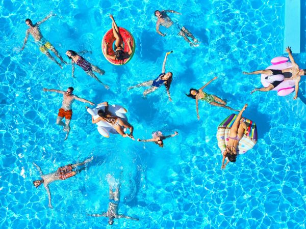 Aerial of friends having party in swimming pool with inflatable flamingo, swan, mattresses. Happy young people relax at luxury resort on sunny day. View from above. Girls in bikini sunbathing in sun.