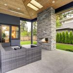 New modern home features a backyard with covered patio accented with stone fireplace, vaulted ceiling with skylights and furnished with gray wicker sofa placed on concrete floor. Northwest, USA