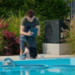 A pool maintenance technician in a green t-shirt and overalls is kneeling by the edge of a swimming pool, closely reviewing data on a tablet. The scene is set in a well-maintained outdoor area surrounded by tall ornamental grasses and plants, with two large chemical containers placed near the pool. The technician is focused on ensuring the pool's water quality, emphasizing the importance of professional upkeep in maintaining a clean and safe swimming environment.