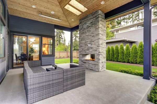 New modern home features a backyard with covered patio accented with stone fireplace, vaulted ceiling with skylights and furnished with gray wicker sofa placed on concrete floor. Northwest, USA